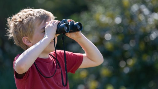 A young boy wearing a red t-shirt looking through a pair of binoculars and smiling